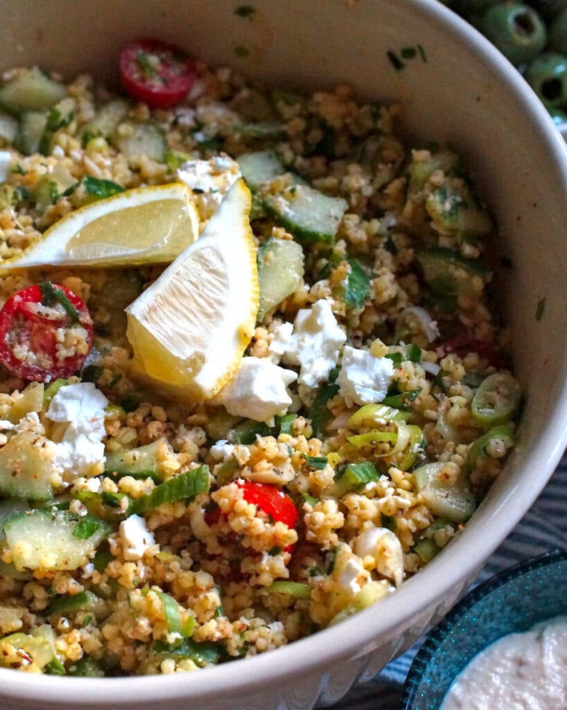 A large bowl of summery millet tabbouleh. Tiny golden millet grains, chopped cucumbers, tomatoes and spring onions are topped with crumbled feta and garnished with lemon wedges.