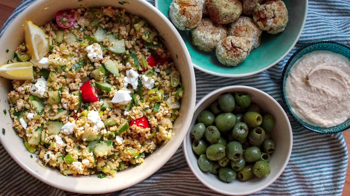Millet tabbouleh served in a large ceramic bowl, sitting on a green striped tea towel. Next to the large bowl are several smaller bowls, filled with olives, white bean hummus and homemade baked falafel.
