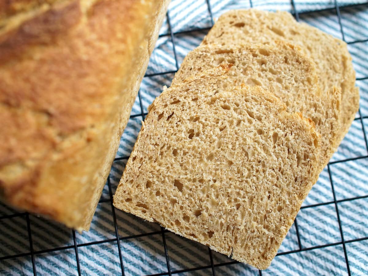Slices of homemade kamut sourdough sandwich bread are stacked on a black wire rack on top of a green and white striped tea towel, next to the golden brown loaf. The crumb is even and soft.