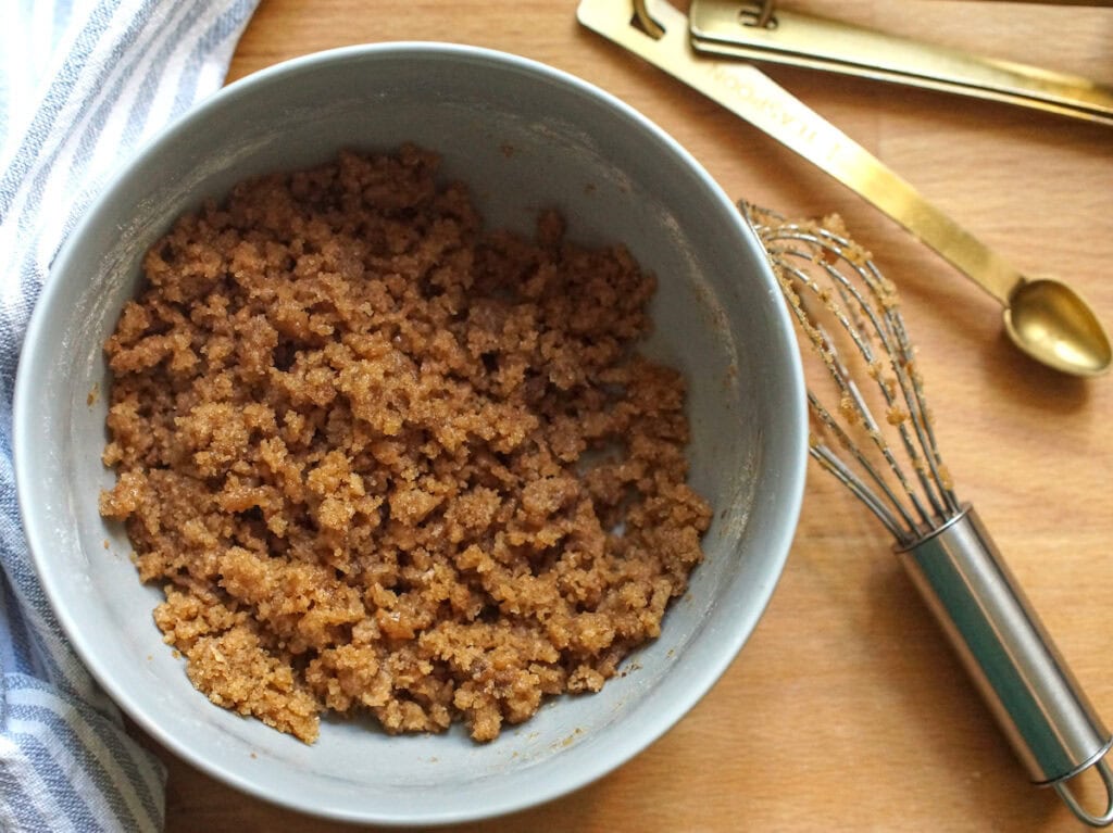 Cinnamon-spiked crumble topping has been prepped in a pale blue bowl, surrounded by a small whisk, golden measuring spoons and a striped tea towel.