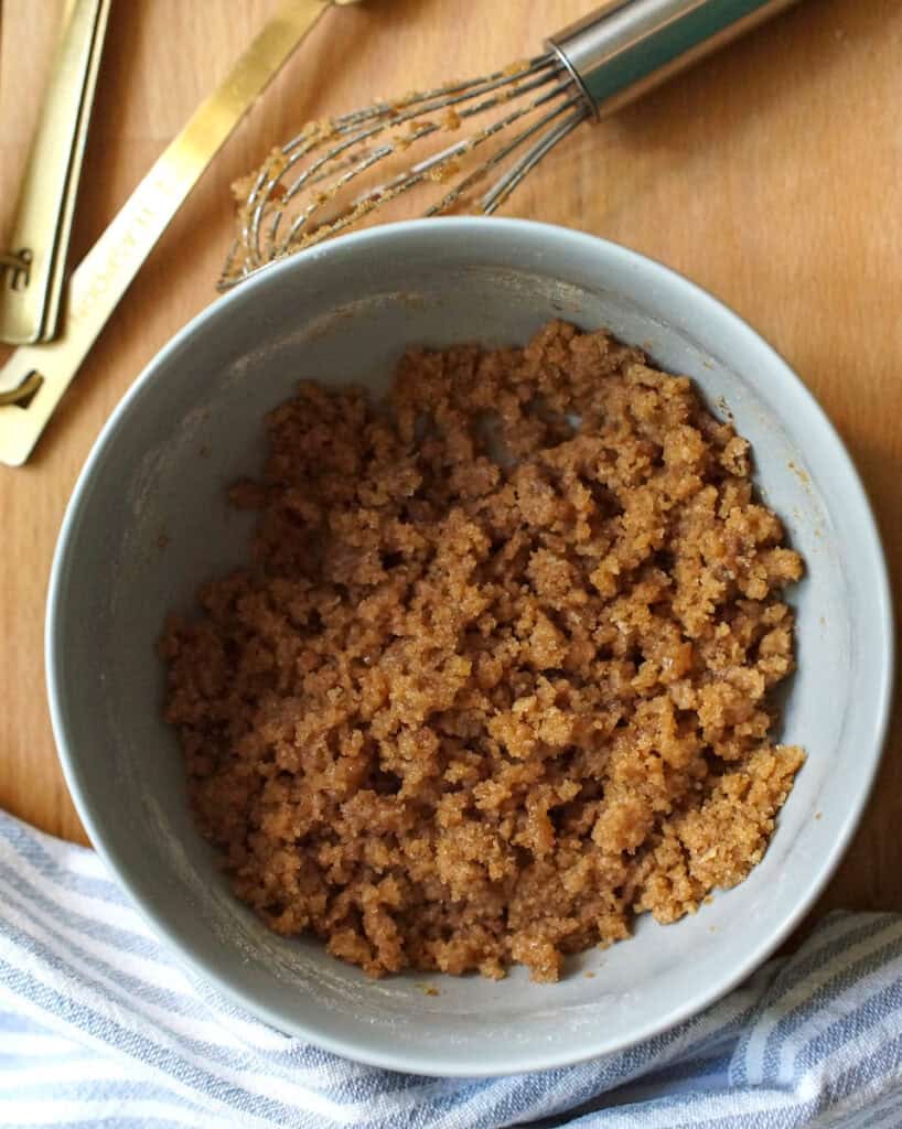 Cinnamon-spiked crumble topping has been prepped in a pale blue bowl, surrounded by a small whisk, golden measuring spoons and a striped tea towel.