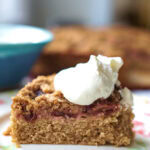 A piece of cardamom-spiced spelt damson buckle is served on a floral plate, topped with bright white whipped cream.
