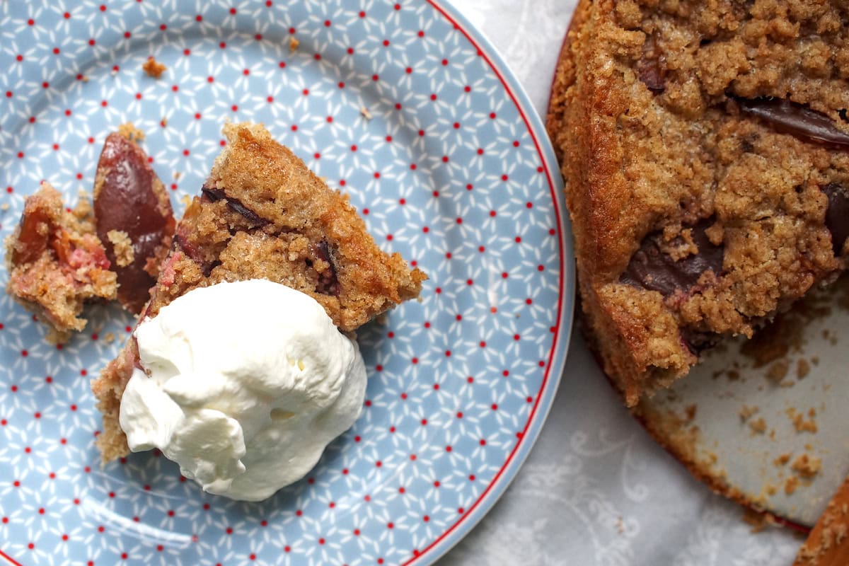 A generous slice of cardamom-spiced spelt damson buckle is served with a dollop of whipped cream on a blue plate with a geometric pattern,