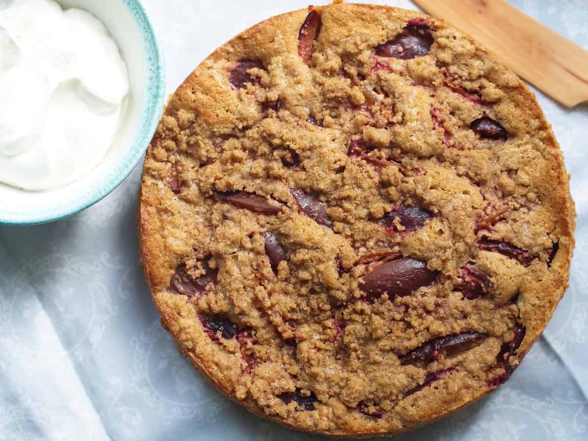 Overhead shot of golden brown spelt damson buckle, sitting on a light grey fabric, next to a bowl of gleaming white whipped cream.
