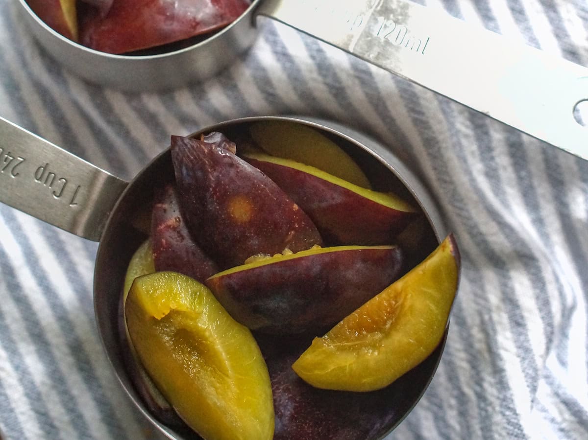 Sliced damson plums in stainless steel measuring cups are sitting on pale blue and white striped tea towel.