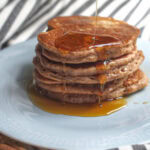 A stack of pancakes with maple syrup being poured from above, dripping down the sides and pooling at the bottom of the plate. Cinnamon sticks and whole einkorn berries are scattered on op of the black and white striped tea towel that's lining the background.