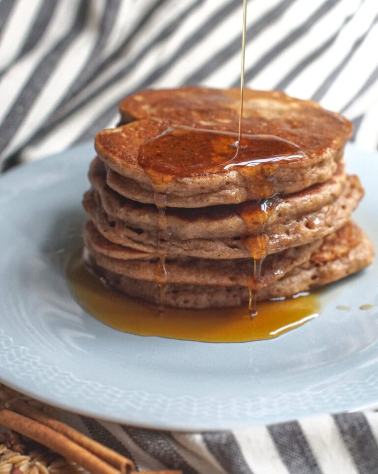 A stack of pancakes with maple syrup being poured from above, dripping down the sides and pooling at the bottom of the plate. Cinnamon sticks and whole einkorn berries are scattered on op of the black and white striped tea towel that's lining the background.