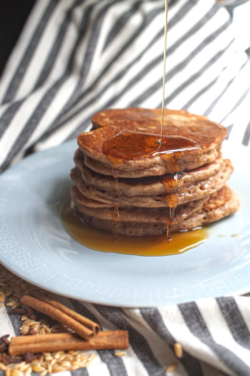 A stack of pancakes with maple syrup being poured from above, dripping down the sides and pooling at the bottom of the plate. Cinnamon sticks and whole einkorn berries are scattered on op of the black and white striped tea towel that's lining the background.