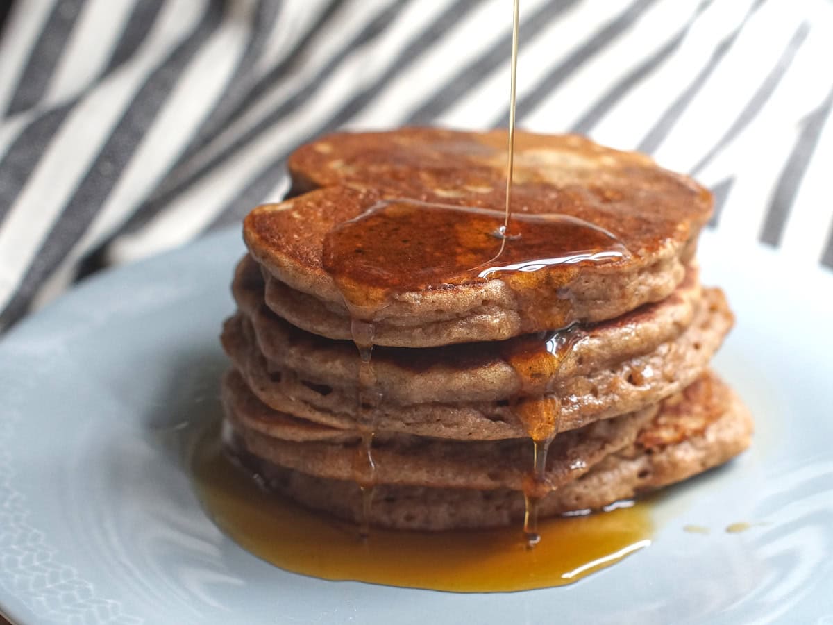 A stack of pancakes with maple syrup being poured from above, dripping down the sides and pooling at the bottom of the plate.