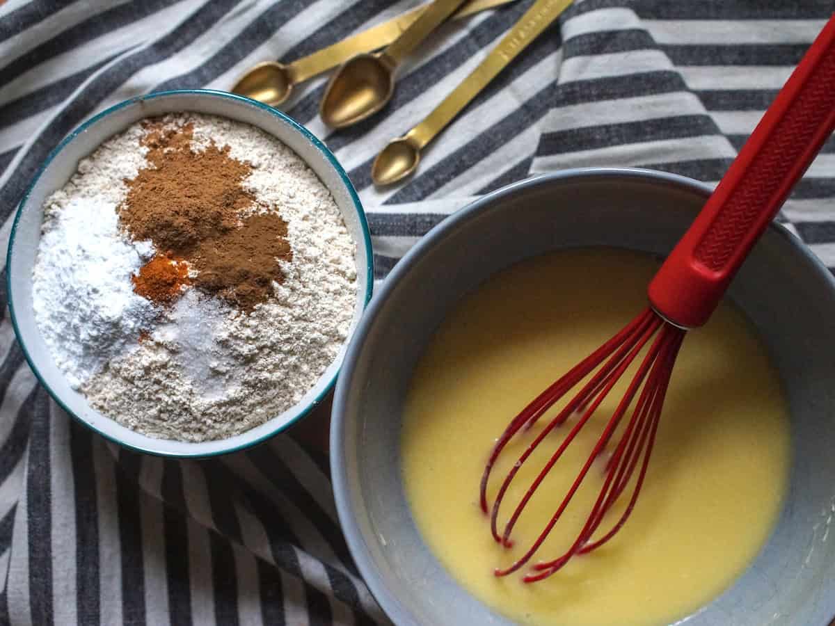 A small bowl filled with freshly milled einkorn flour and dark brown spices, sitting next to a larger bowl, filled with mixed wet ingredients. Both bowls are laid out on a moody, black and white striped tea towel, next to a set of golden measuring spoons.