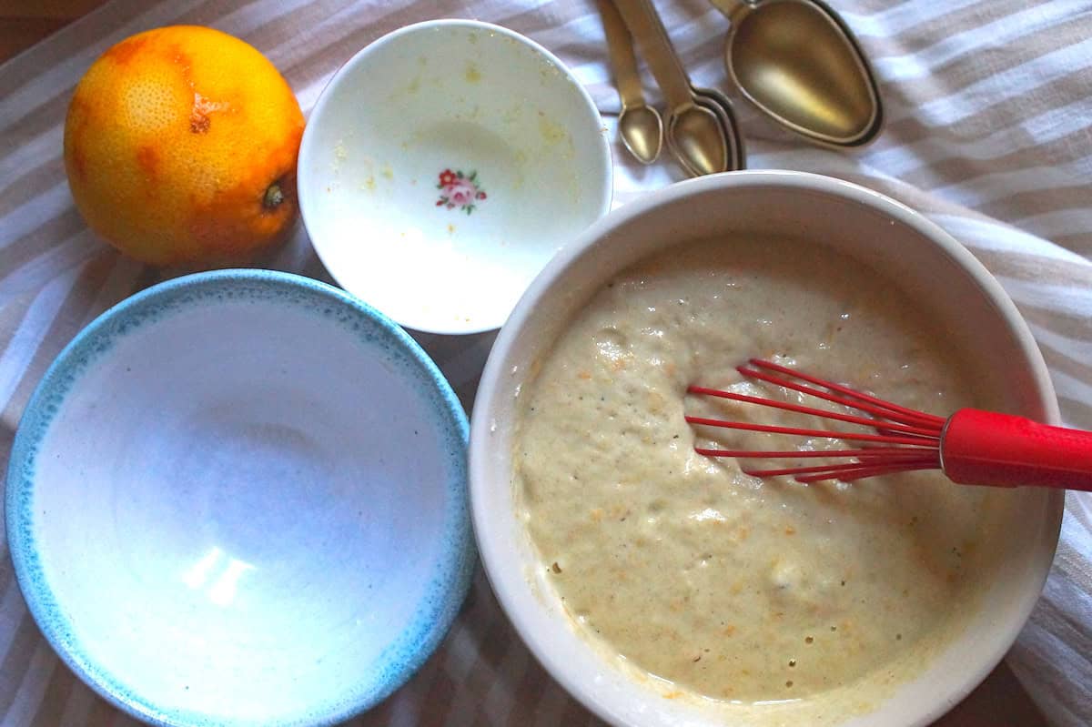 Ingredients for gluten-free sourdough pancakes laid out on a striped tea towel, including two empty bowls for dry ingredients, and one bowl filled with the mixedpancake batter. A zested orange and gold measuring spoons sit beside them.