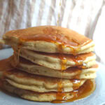 A thick and tall stack of gluten-free sourdough discard pancakes on a pale blue plate. Maple syrup is poured from above, flowing off the sides and pooling at the bottom.