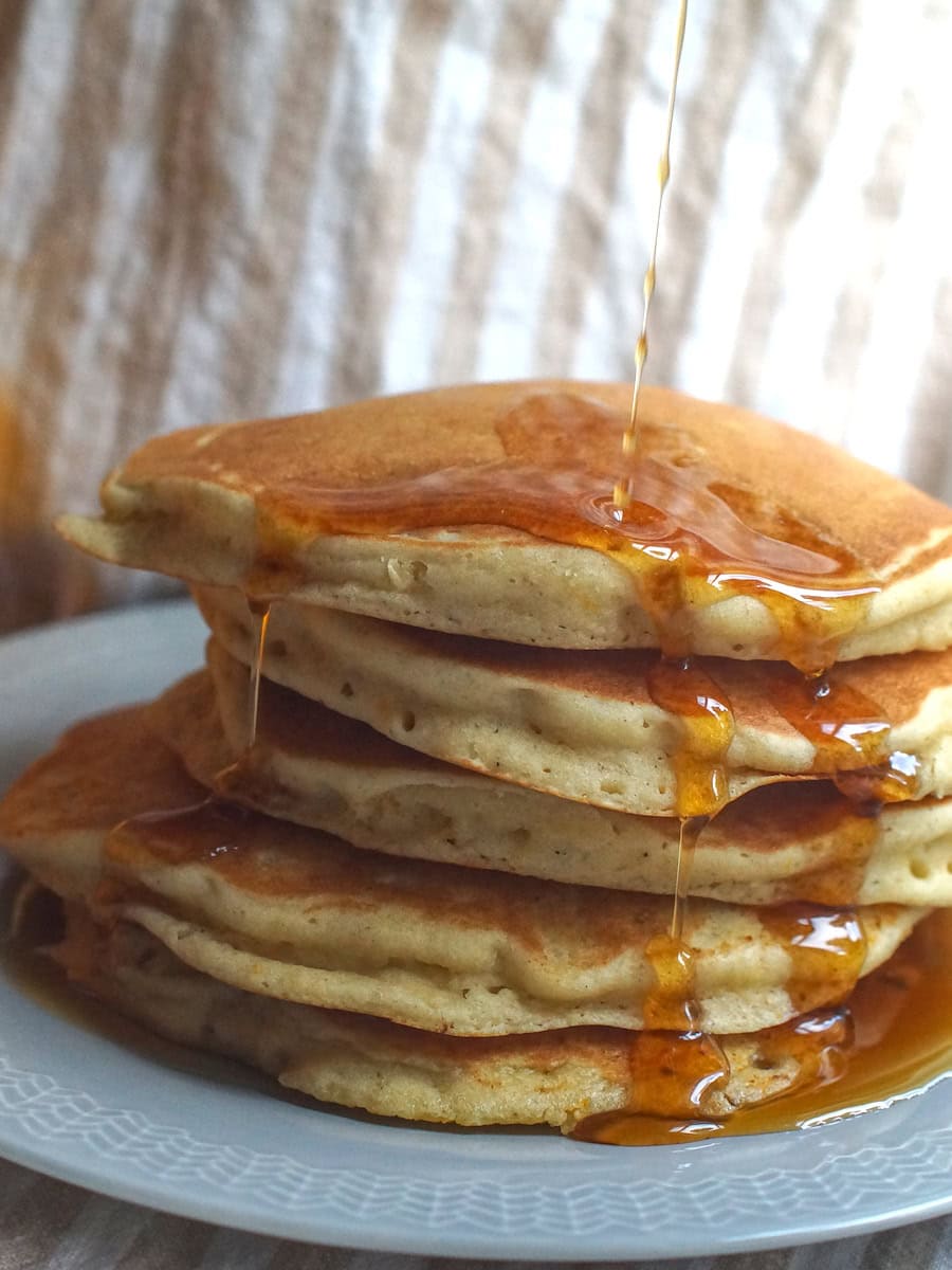 A thick and tall stack of gluten-free sourdough discard pancakes on a pale blue plate. Maple syrup is poured from above, flowing off the sides and pooling at the bottom.