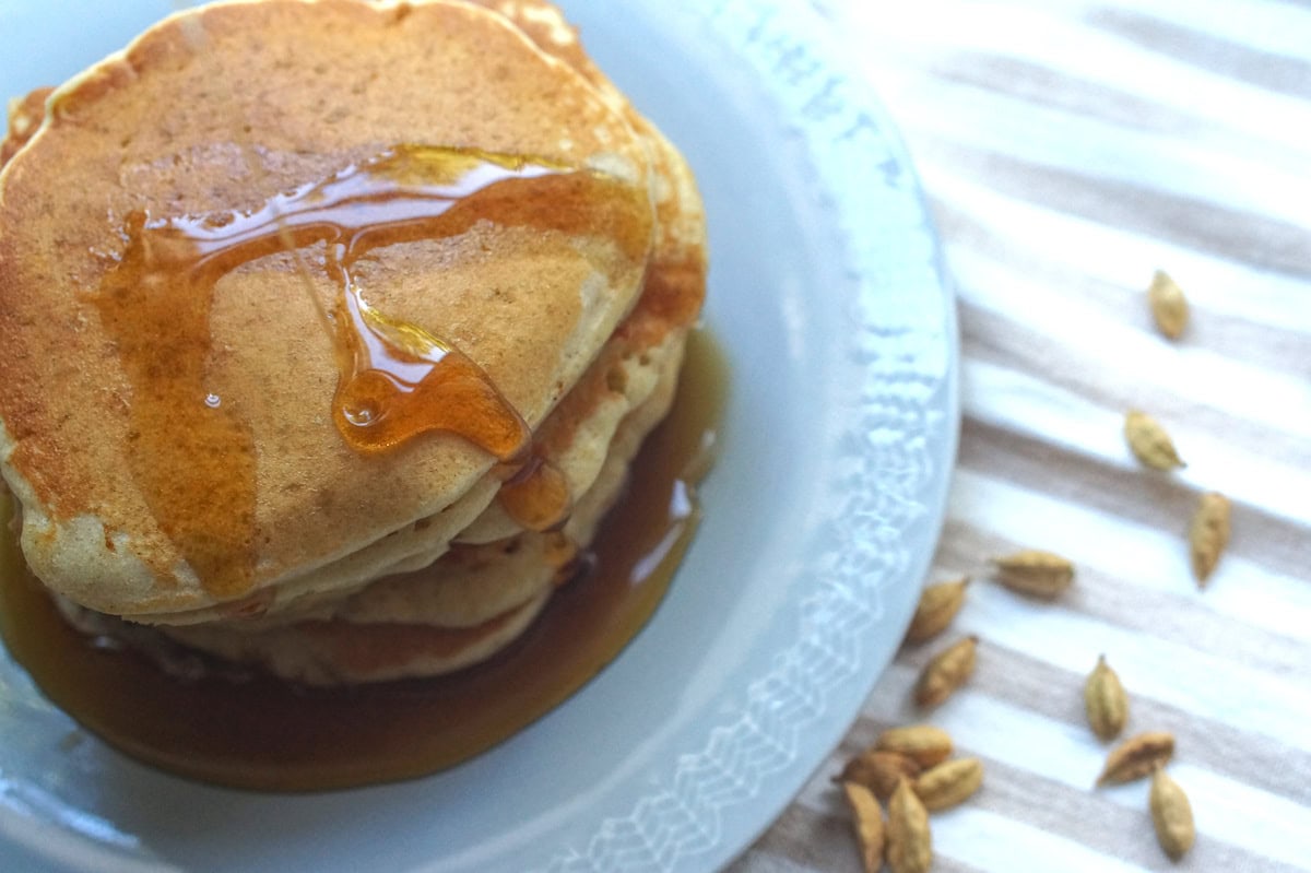 Overhead view of a stack of gluten-free sourdough pancakes with a maple syrup flowing off the sides, pooling at the bottom of the plate. a handful of whole cardamom pots is scattered across the striped tea towel beneath the plate.