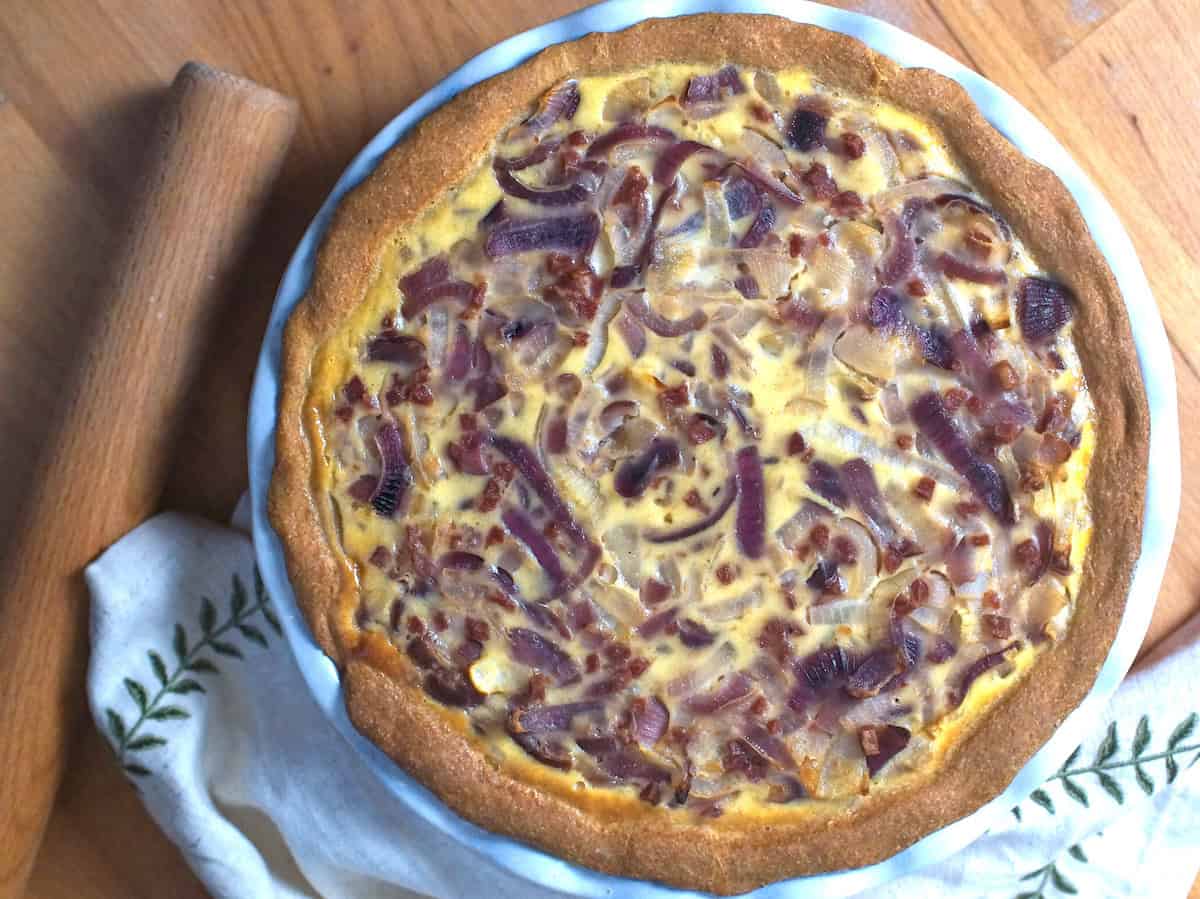 Overhead shot of a German Zwiebelkuchen (onion pie): a golden pie with a browned, bready crust, filled with ribbons of red and yellow onions, bacon bits, and a golden custard filling. The pie sits on a wooden surface, accompanied by an embroidered linen napkin and a wooden rolling pin.
