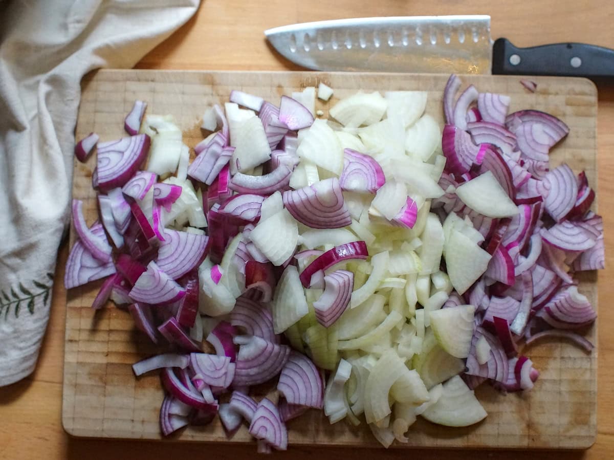 A large pile of chopped red and yellow onions on a wooden chopping board, next to a linen napkin and a large chefs knife.