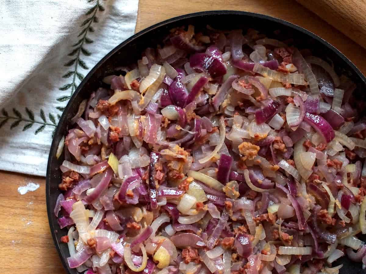 A large frying pan filled with softened red and yellow onions and brown bacon bits sits on a wooden counter, next to an embroidered linen napkin and a wooden rolling pin.