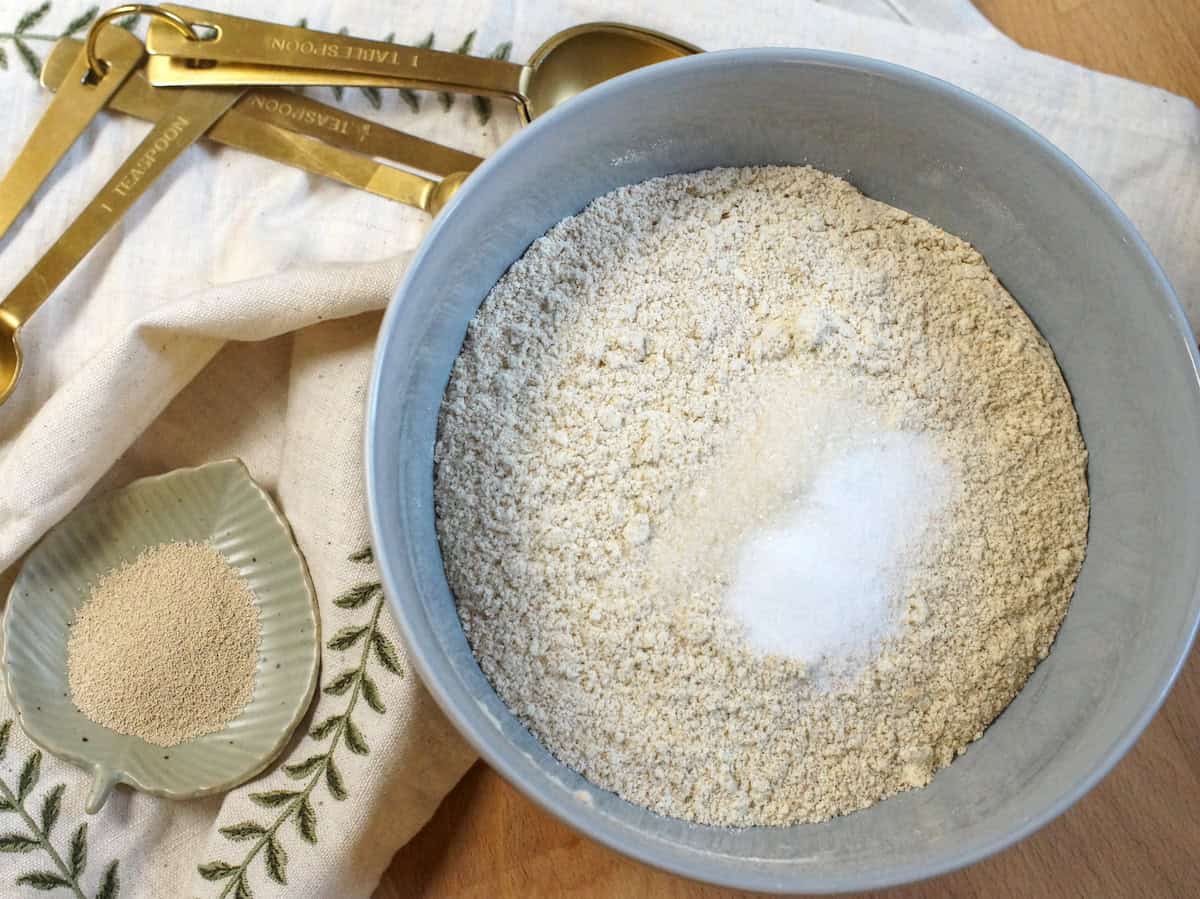 A pale blue ceramic bowl filled with freshly milled soft wheat flour, salt, and sugar. The bowl sits next to an embroidered linen napkin, golden measuring spoons, and a tiny green dish filled with instant yeast.