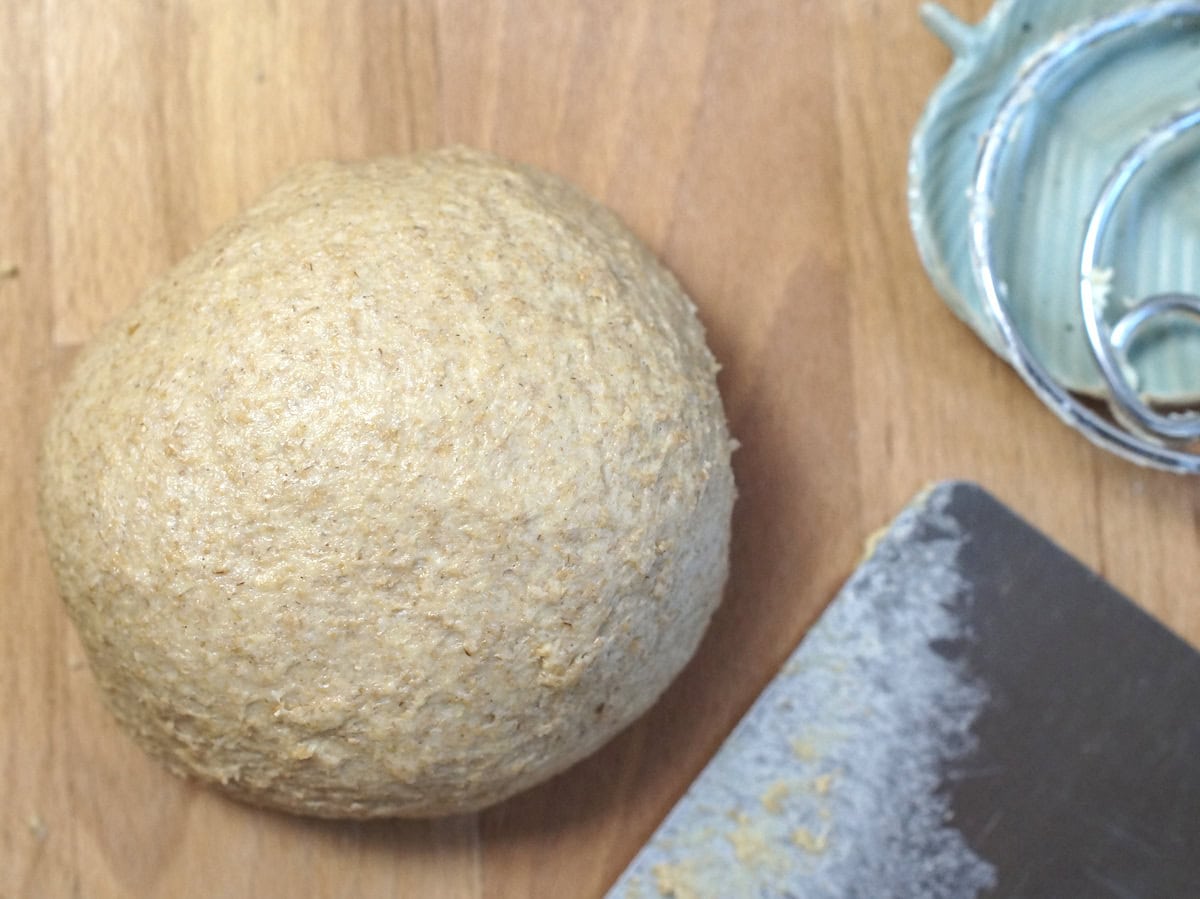 A smooth, shiny ball of dough sits on a wooden kitchen counter, next to a sticky bench scraper.