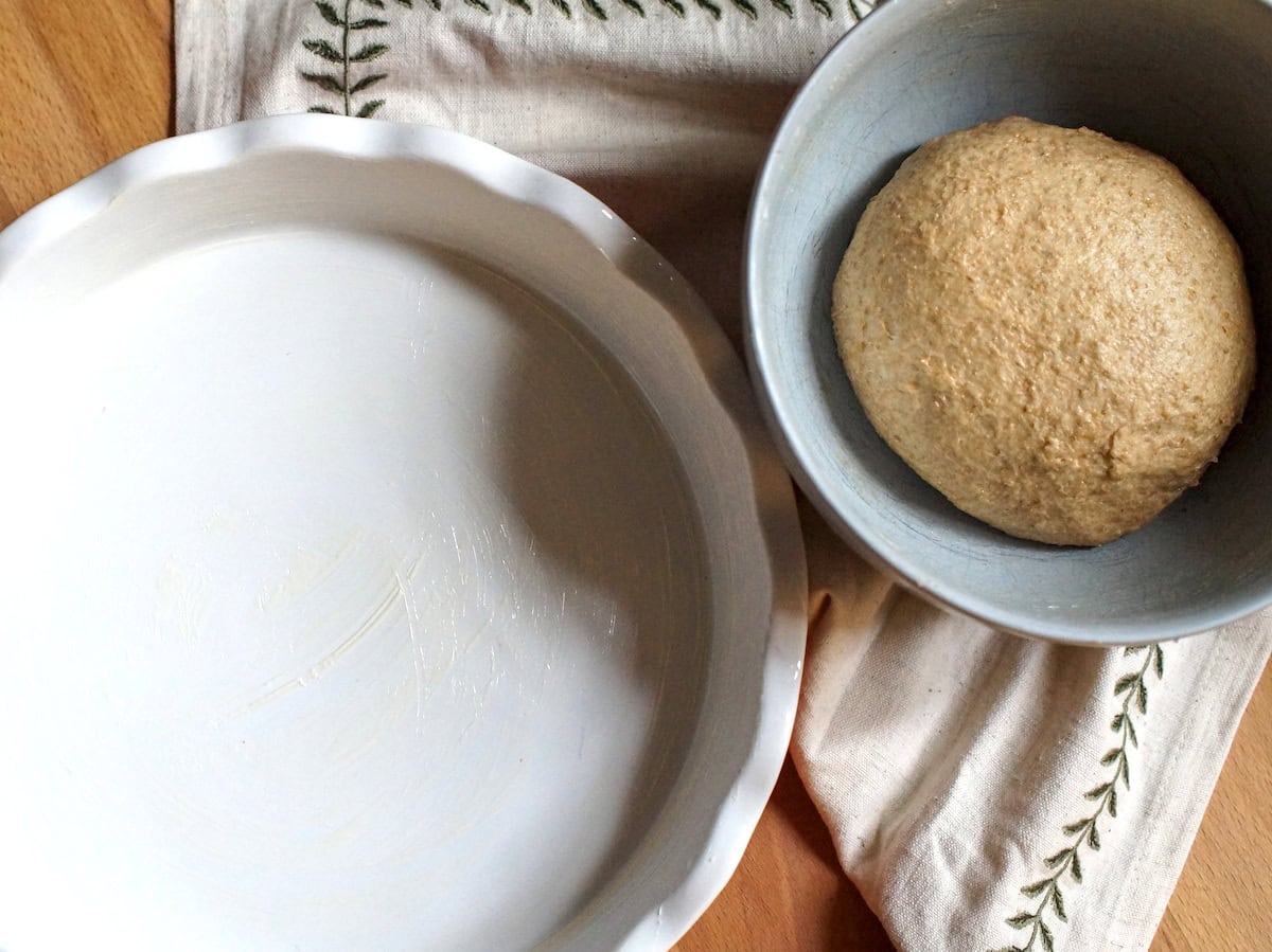 A greased 9-inch pie dish next to a pale blue bowl holding a puffy ball of freshly milled soft wheat dough, ready to be rolled out.