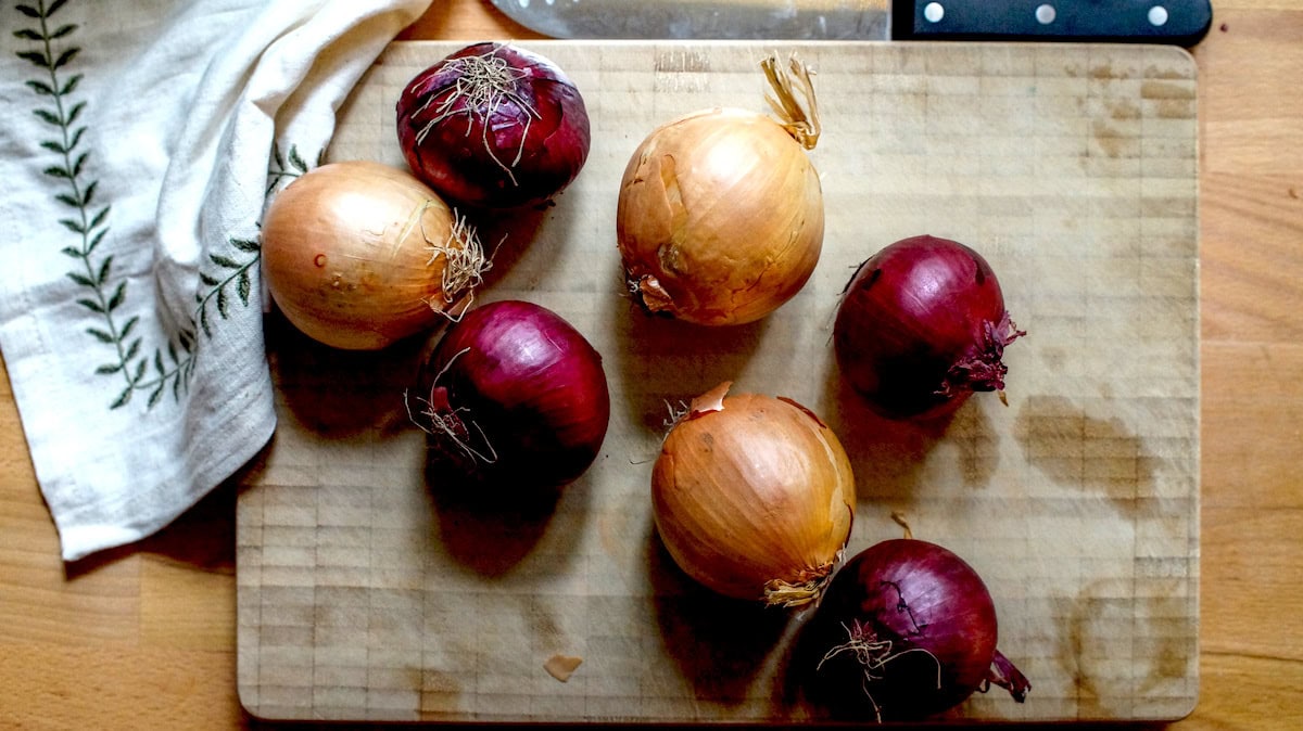 Moody overhead shot of yellow and dark red onions on a stained wooden chopping board, set on a wooden counter beside an embroidered linen napkin and a chef’s knife.