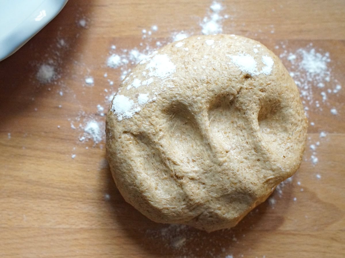 A puffy ball of freshly milled flour dough on a flour-dusted wooden counter. Finger imprints on the dough’s surface show how soft and pliable it is.
