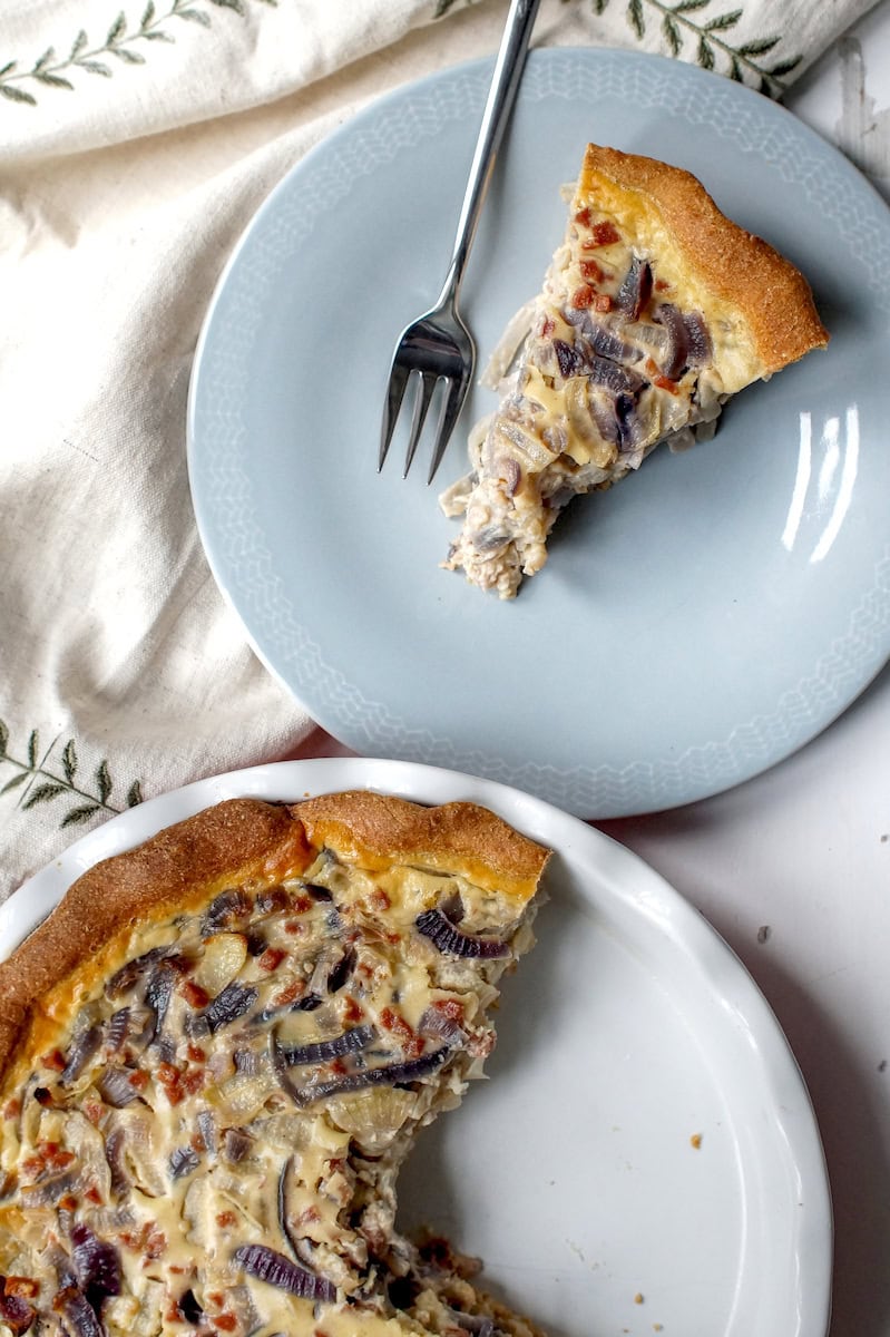 White fluted ceramic pie dish, three-quarters filled with German Zwiebelkuchen (onion pie), sits on a chipped white surface next to a pale blue plate holding a slice of the pie. A linen napkin with green embroidered leaves completes the scene.