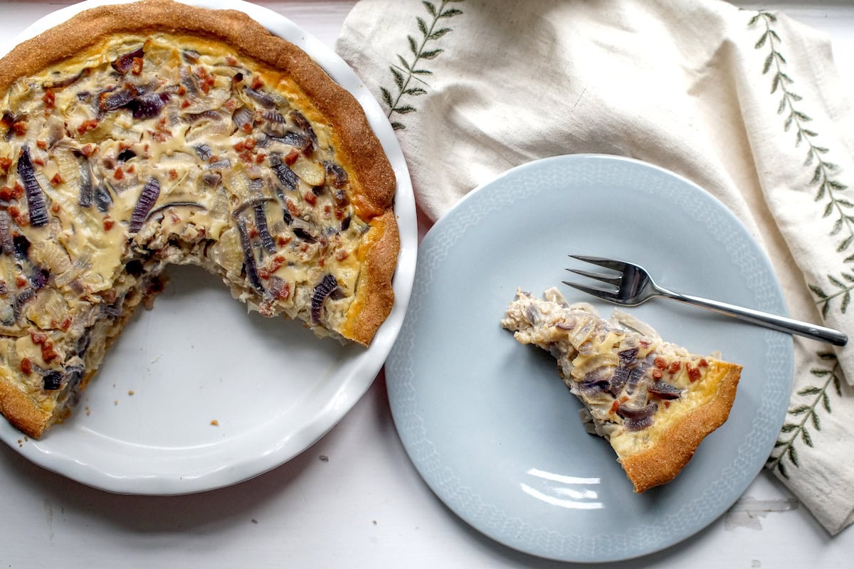 White fluted ceramic pie dish, three-quarters filled with German Zwiebelkuchen (onion pie), sits on a chipped white surface next to a pale blue plate holding a slice of the pie. A linen napkin with green embroidered leaves completes the scene.
