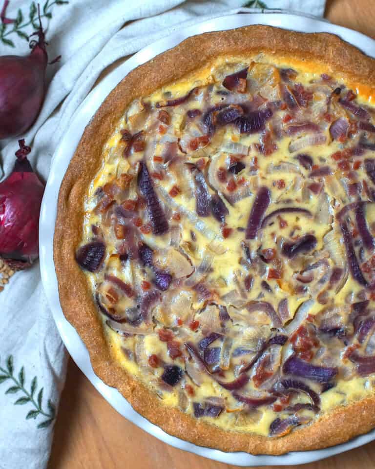 Overhead shot of a German Zwiebelkuchen (onion pie): a golden pie with a browned, bready crust, filled with ribbons of red and yellow onions, bacon bits, and a golden custard filling. The pie sits on a wooden surface, accompanied by an embroidered linen napkin and two purple-red onions.