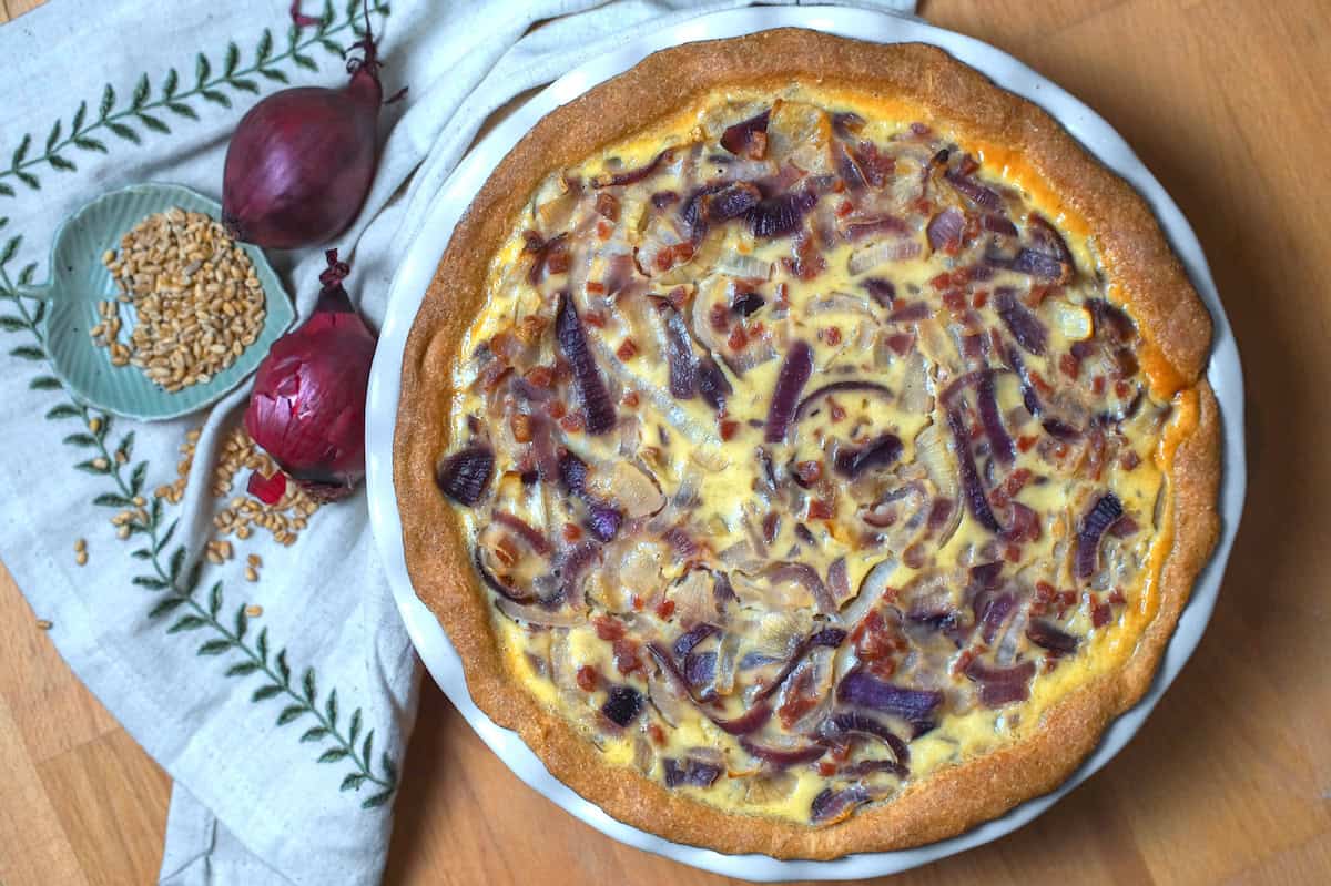 Overhead shot of a German Zwiebelkuchen (onion pie): a golden pie with a browned, bready crust, filled with ribbons of red and yellow onions, bacon bits, and a golden custard filling. The pie sits on a wooden surface, accompanied by an embroidered linen napkin, two purple-red onions, and a tiny green dish filled with whole soft wheat berries.