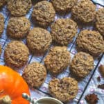 Fresh milled spelt sourdough chocolate chip cookies on a black wire rack sitting on a patterned tea towel. Whole fall spices are scattered on the side, next tu a bright orange pumpkin and a cup of frothy milk tea.