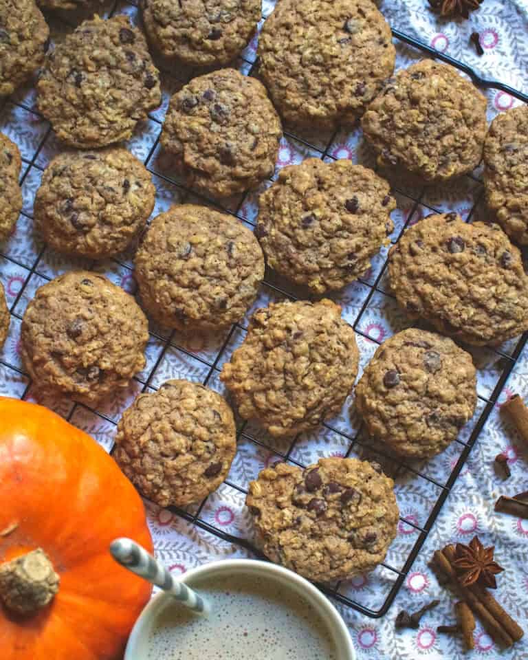 Fresh milled spelt sourdough chocolate chip cookies on a black wire rack sitting on a patterned tea towel. Whole fall spices are scattered on the side, next tu a bright orange pumpkin and a cup of frothy milk tea.