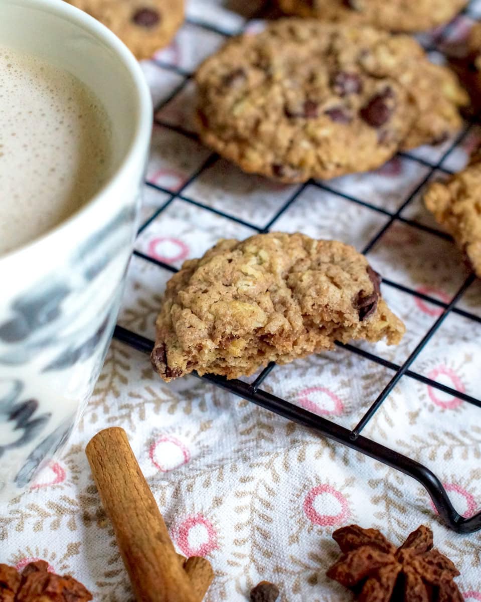 Closeup of half a spelt and oat sourdough chocolate chip cookie, resting on a black wire rack that's sitting o a patterned tea towel. Whole fall spices are visible at the side, next to a lightly grey, floral latte cup filled with foamy white milk. More chocolate chip cookies are cooling off in the background.