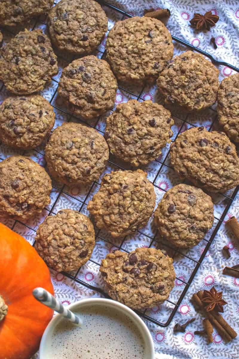 Fresh milled spelt sourdough chocolate chip cookies on a black wire rack sitting on a patterned tea towel. Whole fall spices are scattered on the side, next tu a bright orange pumpkin and a cup of frothy milk tea.