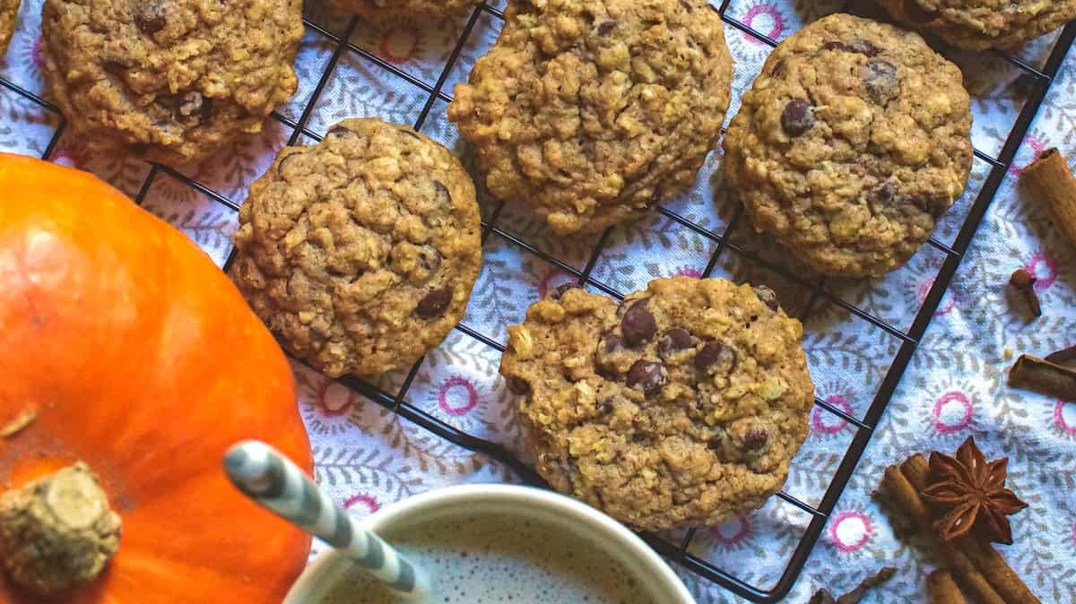 Fresh milled spelt sourdough chocolate chip cookies on a black wire rack sitting on a patterned tea towel. Whole fall spices are scattered on the side, next tu a bright orange pumpkin and a cup of frothy milk tea.
