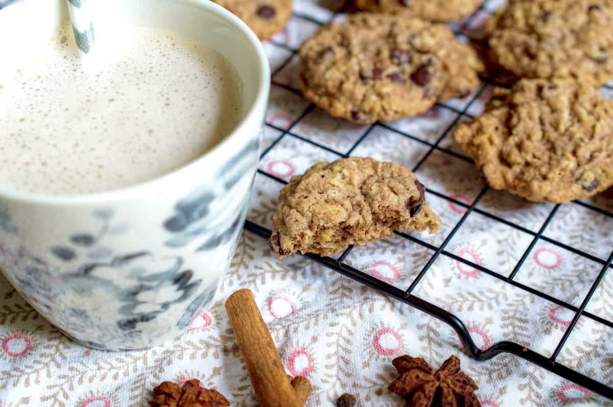 Closeup of half a spelt and oat sourdough chocolate chip cookie, resting on a black wire rack that's sitting o a patterned tea towel. Whole fall spices are visible at the side, next to a lightly grey, floral latte cup filled with foamy white milk. More chocolate chip cookies are cooling off in the background.