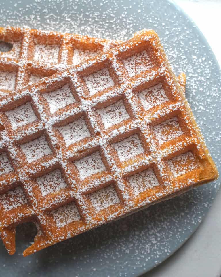 Two crispy, golden sourdough discard waffles, stacked on a pale blue plate, generously dusted with powdered sugar.