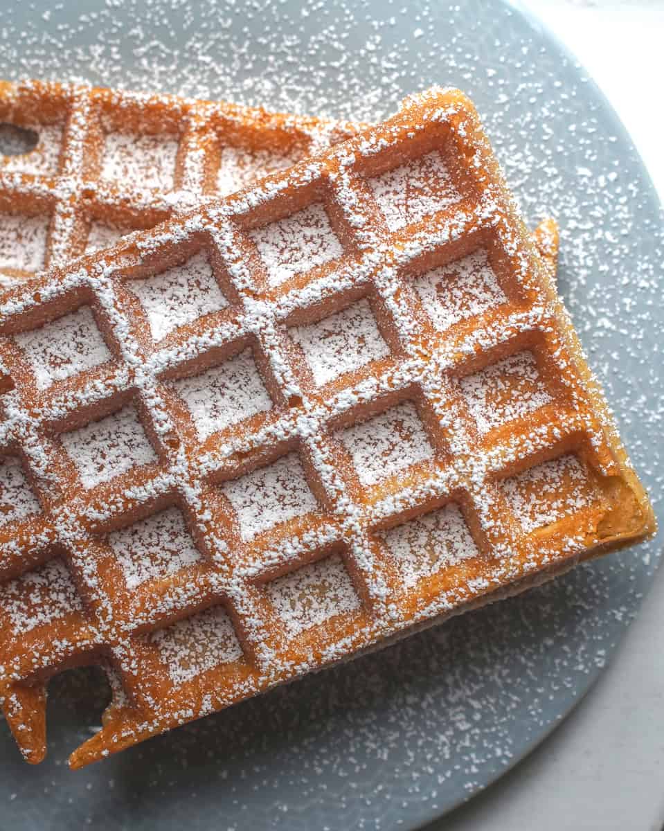 Two crispy, golden sourdough discard waffles, stacked on a pale blue plate, generously dusted with powdered sugar.