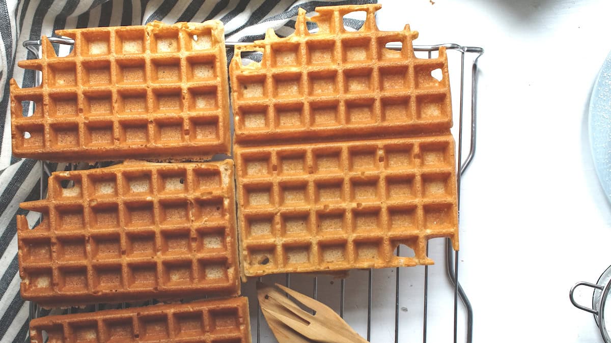 Golden crispy gluten free sourdough waffles sitting on a cooling rack on a striped tea towel, with a pair of wooden fork and knife to the side.