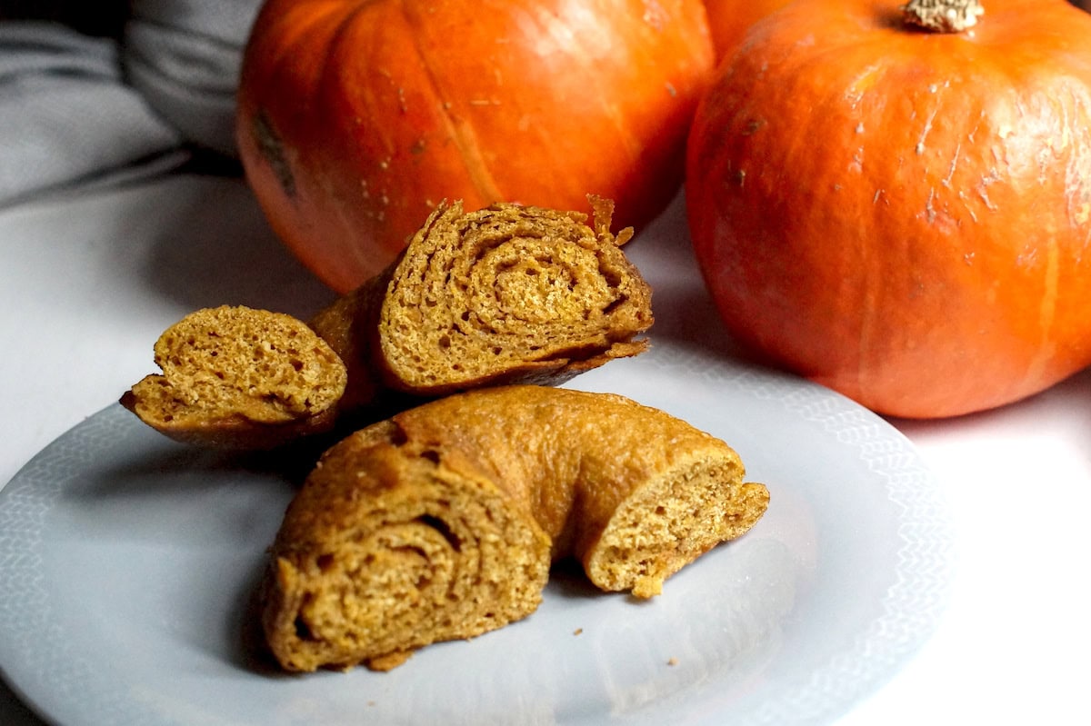One fresh-milled pumpkin sourdough bagel, sitting on a light blue late, next to a pile of bright orange pumpkins. The orange hued whole grain bagel is split in the middle, to expose the dark brown cinnamon sugar swirl filling.