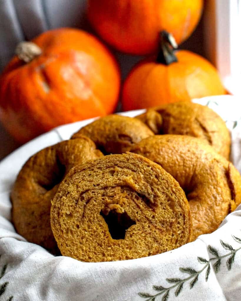 Whole grain fresh-milled pumpkin sourdough bagels served in a basket lined with an embroidered linen cloth. One bagel is split to expose the dark brown cinnamon sugar swirl.