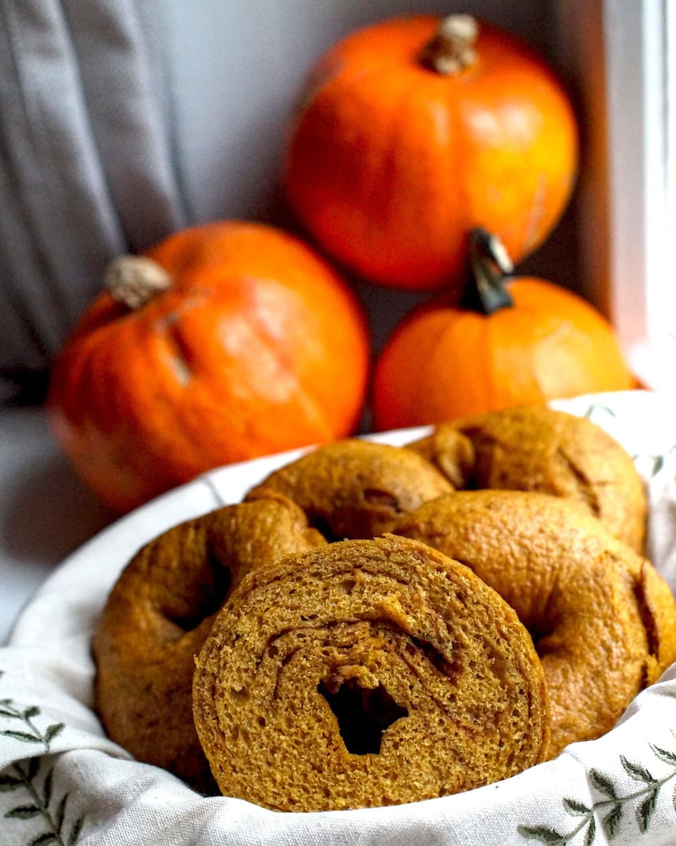 Whole grain fresh-milled pumpkin sourdough bagels served in a basket lined with an embroidered linen cloth. One bagel is split to expose the dark brown cinnamon sugar swirl.