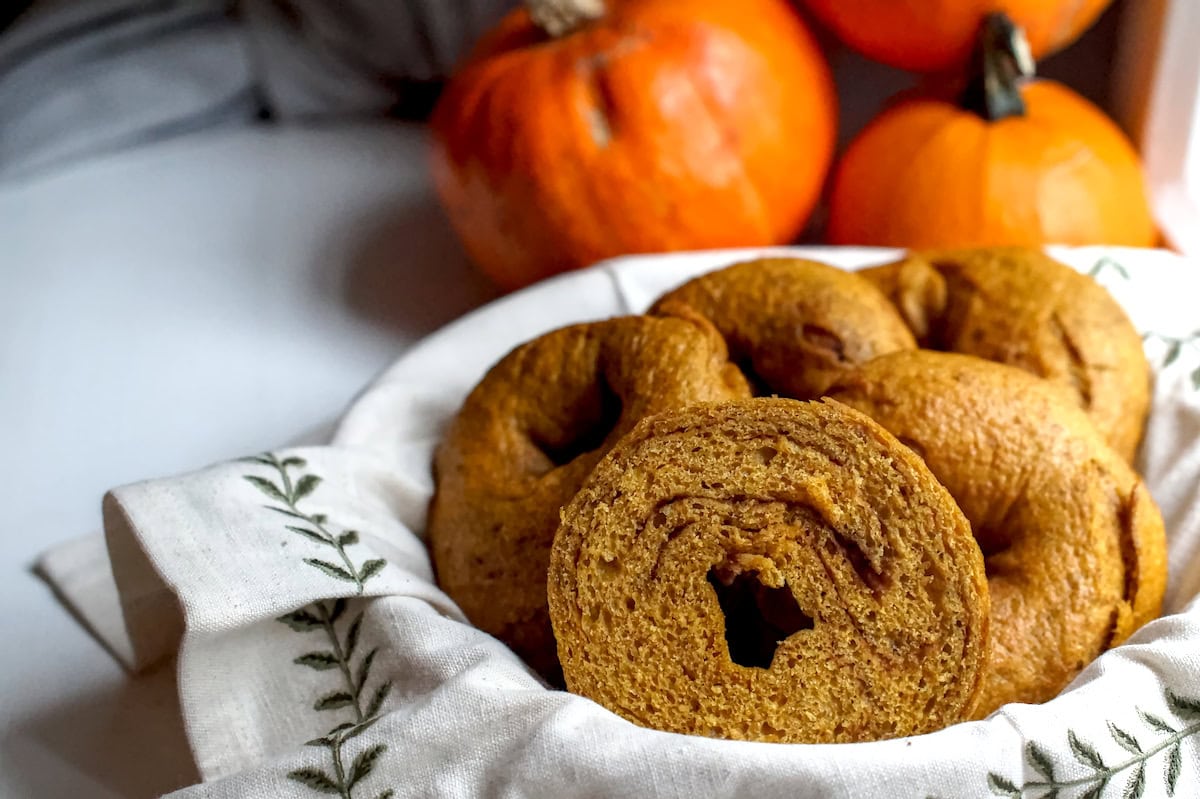 Whole grain fresh-milled pumpkin sourdough bagels served in a basket lined with an embroidered linen cloth. One bagel is split to expose the dark brown cinnamon sugar swirl.