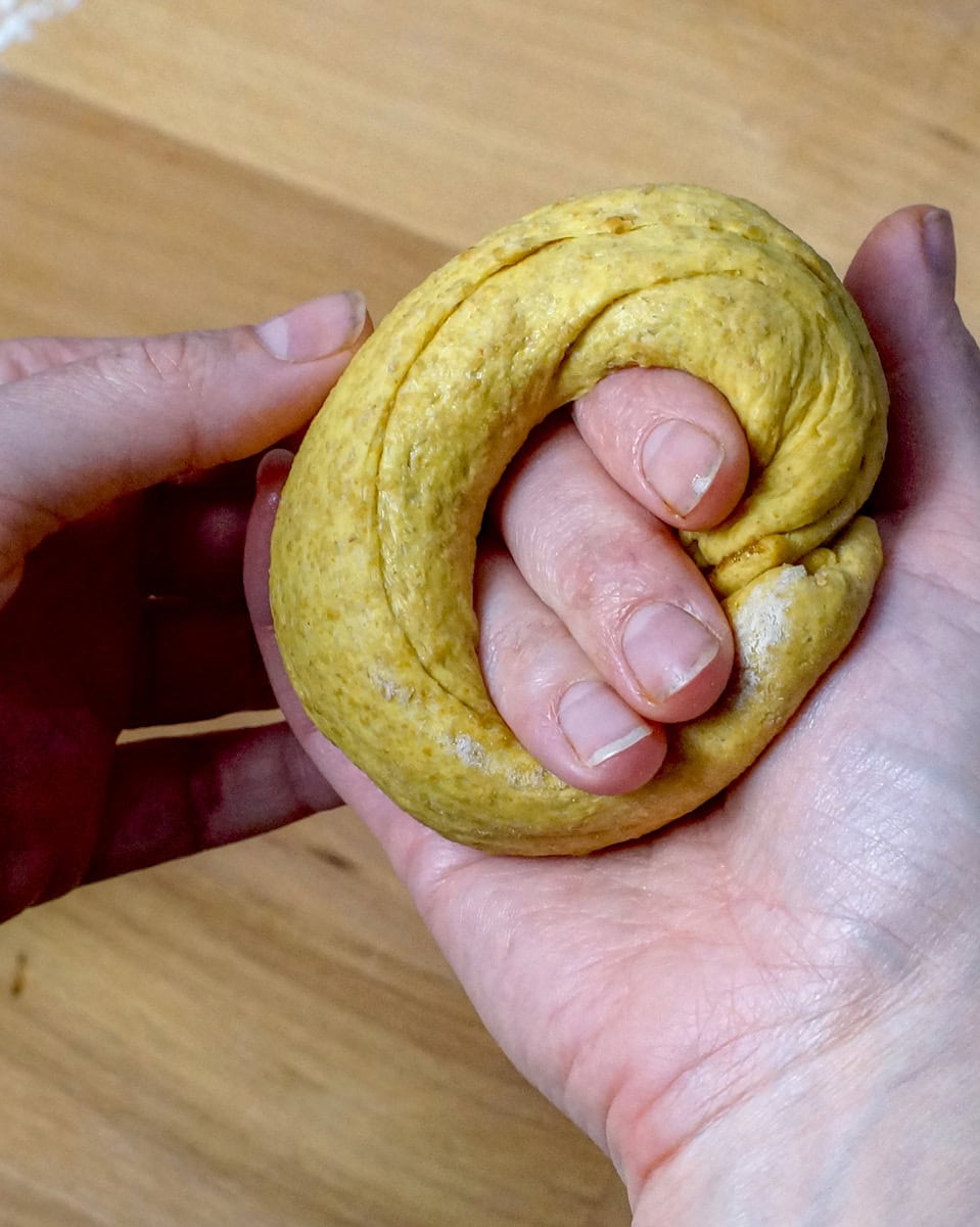 A cinnamon-swirled rope of sourdough pumpkin bagel dough rapped around three fingers of one hand, showing how to pinch together the ends to form a round bagel.