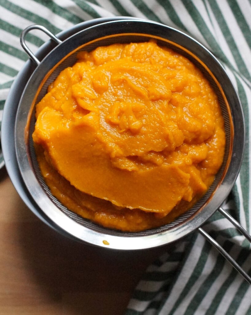 Bright orange roasted pumpkin puree draining in a fine mesh sieve set over a pale blue bowl on a wooden counter with a green and white striped tea towel.