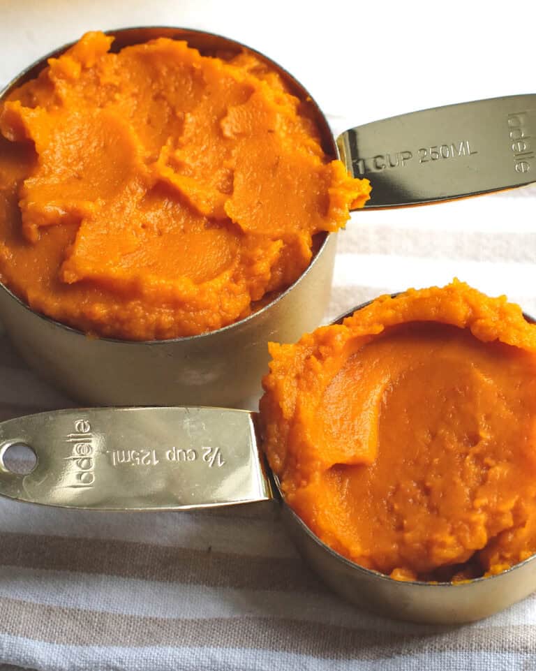 Homemade roasted pumpkin puree in two golden measuring cups, sitting on a tan and white striped tea towel.