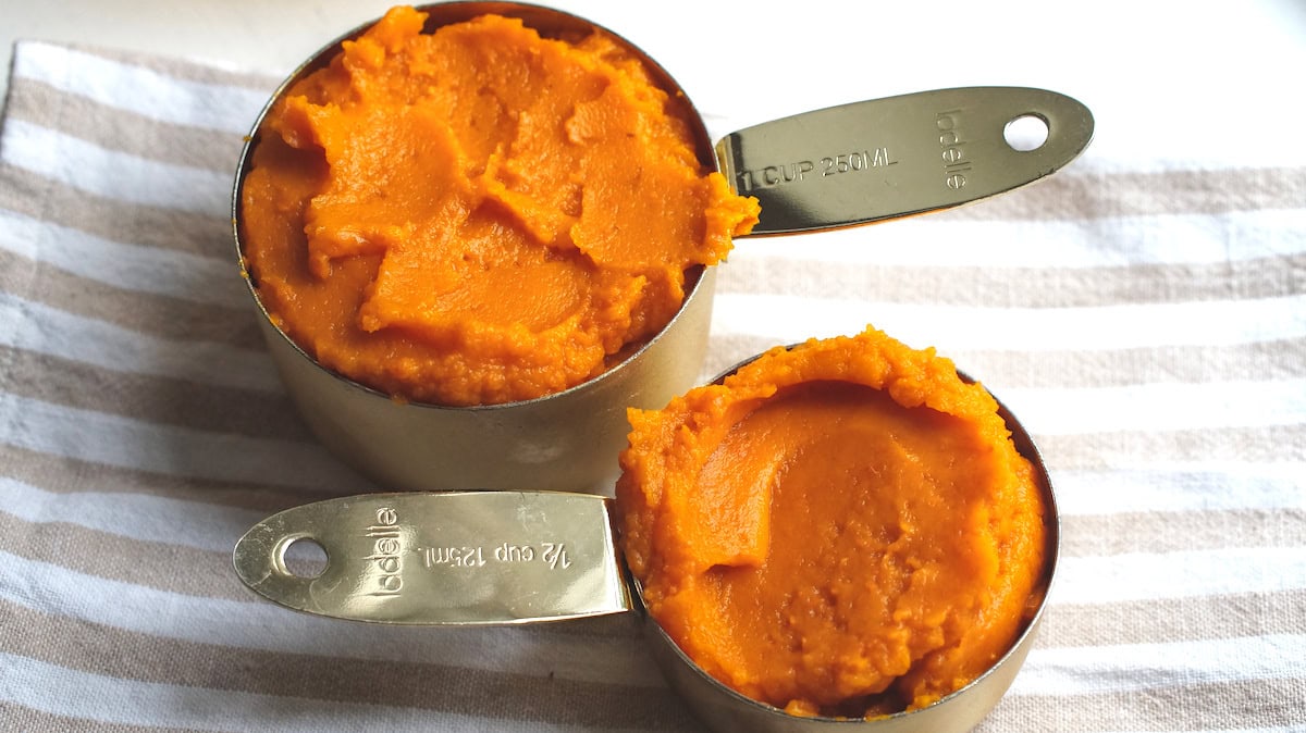 Homemade roasted pumpkin puree in two golden measuring cups, sitting on a tan and white striped tea towel.