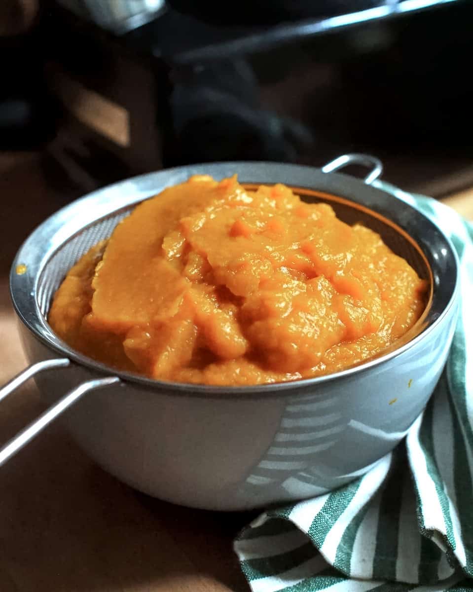 Roasted Pumpkin Puree sitting in a fine mesh sieve over a pale blue bowl, draining after blending in a food processor.