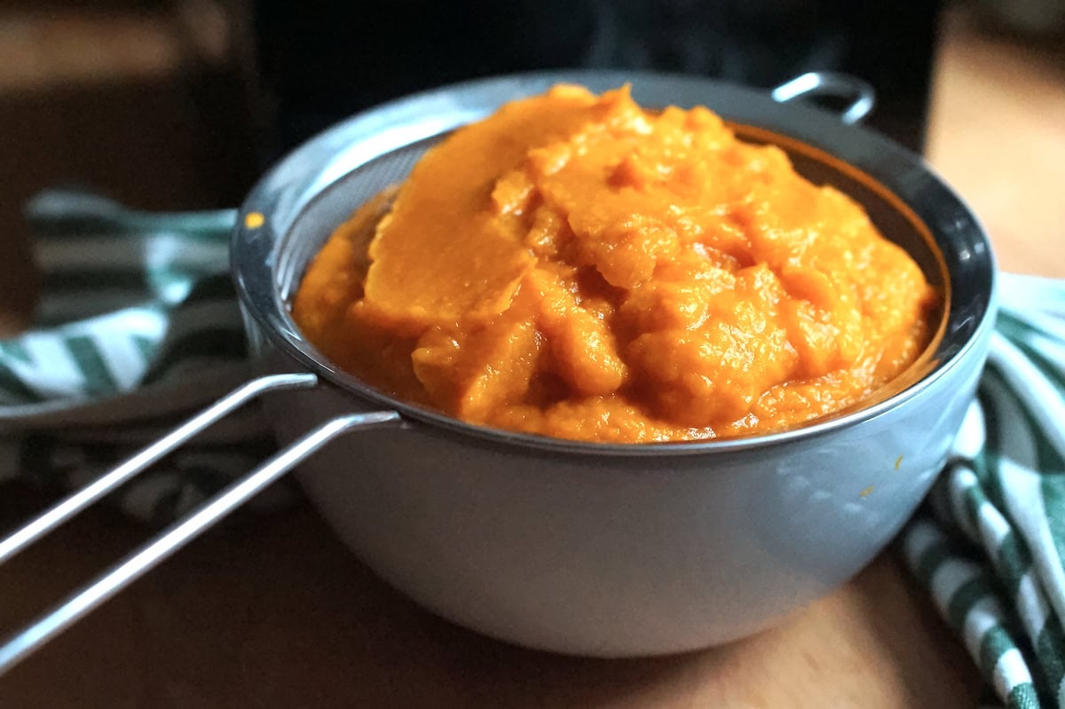 Roasted Pumpkin Puree sitting in a fine mesh sieve over a pale blue bowl, draining after blending in a food processor.