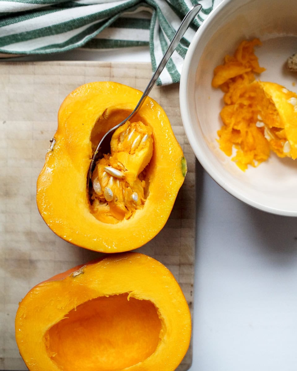 A small orange pumpkin, cut in half on a stained wooden cutting board, with seeds being scooped into a ceramic bowl.